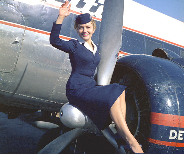 Delta stewardess sitting on DC-3 propeller waving. Wearing 1957-59 Winter Uniform.